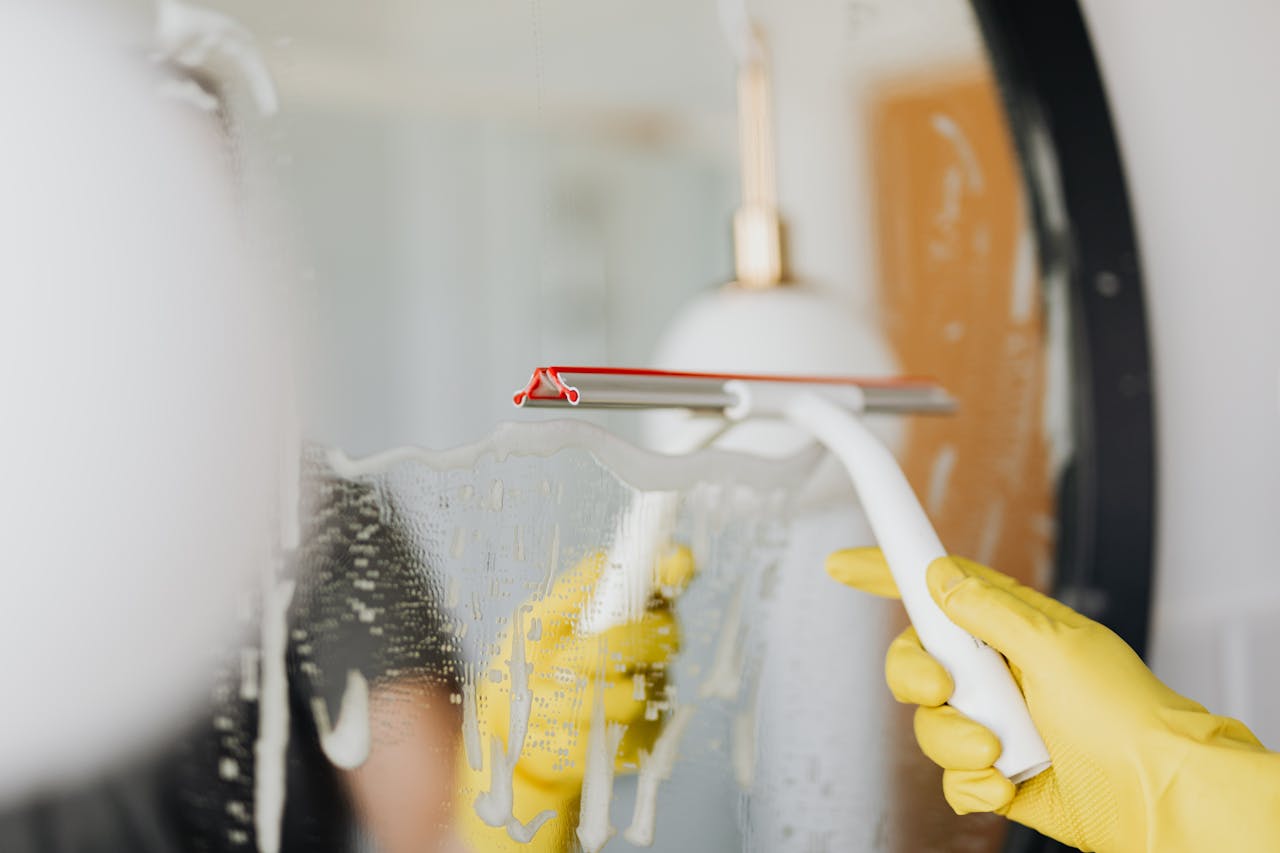 gallery-01 Anonymous person in yellow rubber gloves wiping water by squeegee from mirror in bathroom